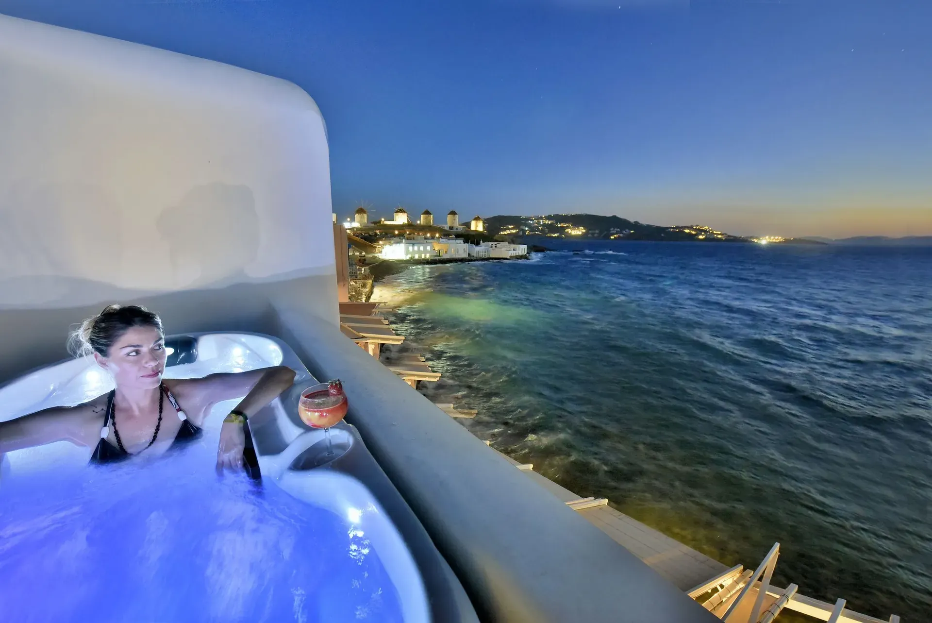 Guest in the lit Jacuzzi at dusk with the windmills across the bay